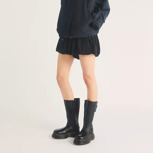 A model from the waist down wearing black mid-calf boots with a distinctive gold-colored stitch detail around the sole, paired with a black pleated mini skirt and a dark navy jacket, against a plain light background.