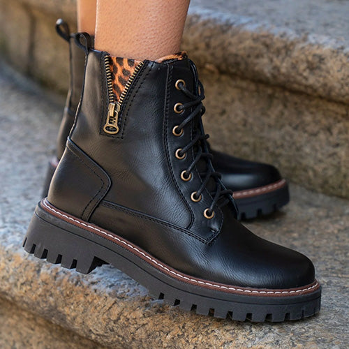 Black leather lace-up ankle boots with a side zipper, gold-tone eyelets, and chunky lug soles, shown worn on stone steps.