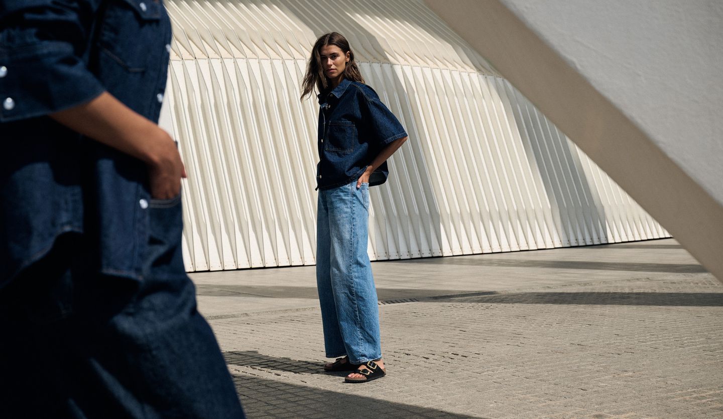 Woman wearing denim shirt and jeans with Birkenstock Arizona Big Buckle black sandals standing on stone pavement, showcasing casual summer style