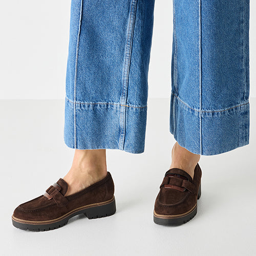Brown suede loafers worn by a person with blue jeans on a white background 