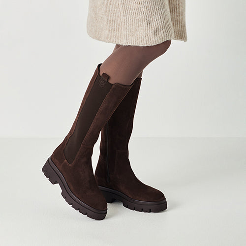 Brown knee-high suede boots worn with a beige skirt on a light gray background