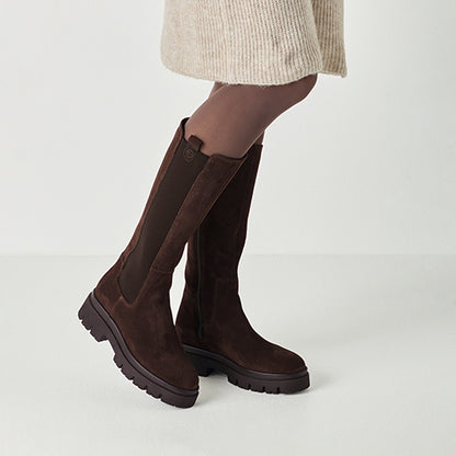 Brown knee-high suede boots worn with a beige skirt on a light gray background