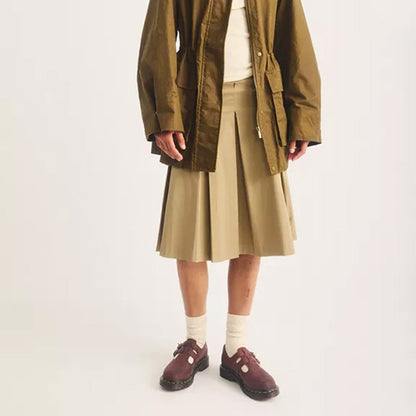 A model is shown from the chest down wearing an olive green utility jacket over a cream top, a beige pleated midi skirt, cream socks, and burgundy T-strap loafers, against a plain off-white studio background.
