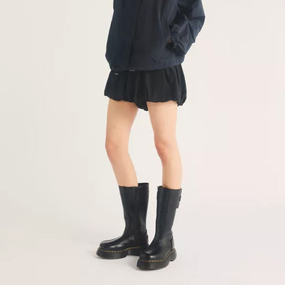 A model from the waist down wearing black mid-calf boots with a distinctive gold-colored stitch detail around the sole, paired with a black pleated mini skirt and a dark navy jacket, against a plain light background.