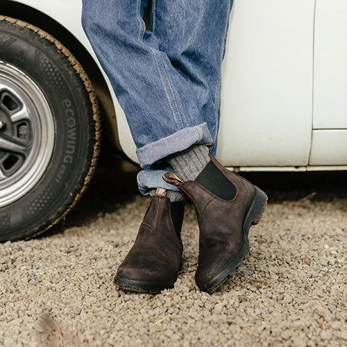 A person wearing dark brown suede Chelsea boots with gray knit socks and blue jeans, standing on gravel next to a white vehicle.