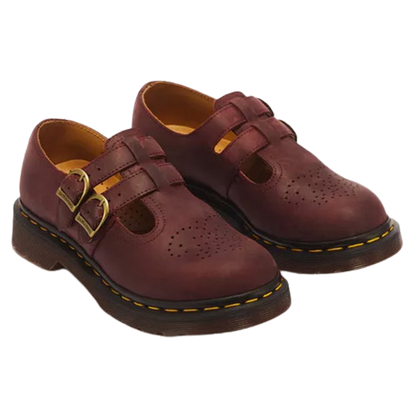 A pair of Dr. Martens double-strap leather shoes in burgundy, featuring gold-tone buckle details and classic yellow welt stitching on a white background.