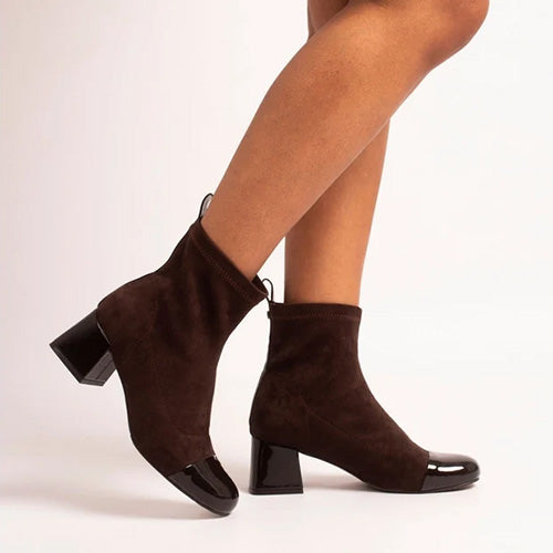 A pair of dark brown suede ankle boots with glossy black patent leather toe caps and block heels, shown on a model&