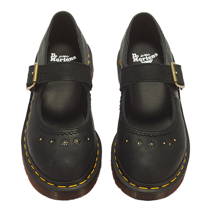 A pair of black Dr. Martens Mary Jane boots with gold-tone buckle straps, floral perforations, and signature yellow stitching on a white background.