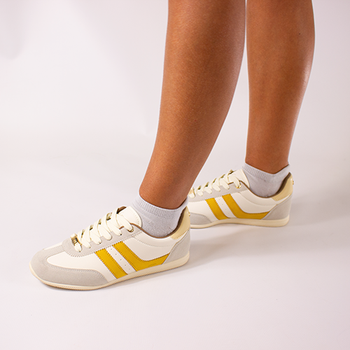 White trainers with yellow stripes worn by a person on a light background