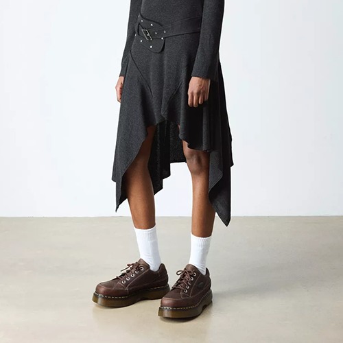Person wearing a dark gray dress with an asymmetrical hem, white socks, and brown buzz shoes on a plain background.