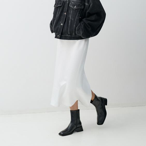 Person wearing a black denim jacket, white skirt, and black shoes on a white background