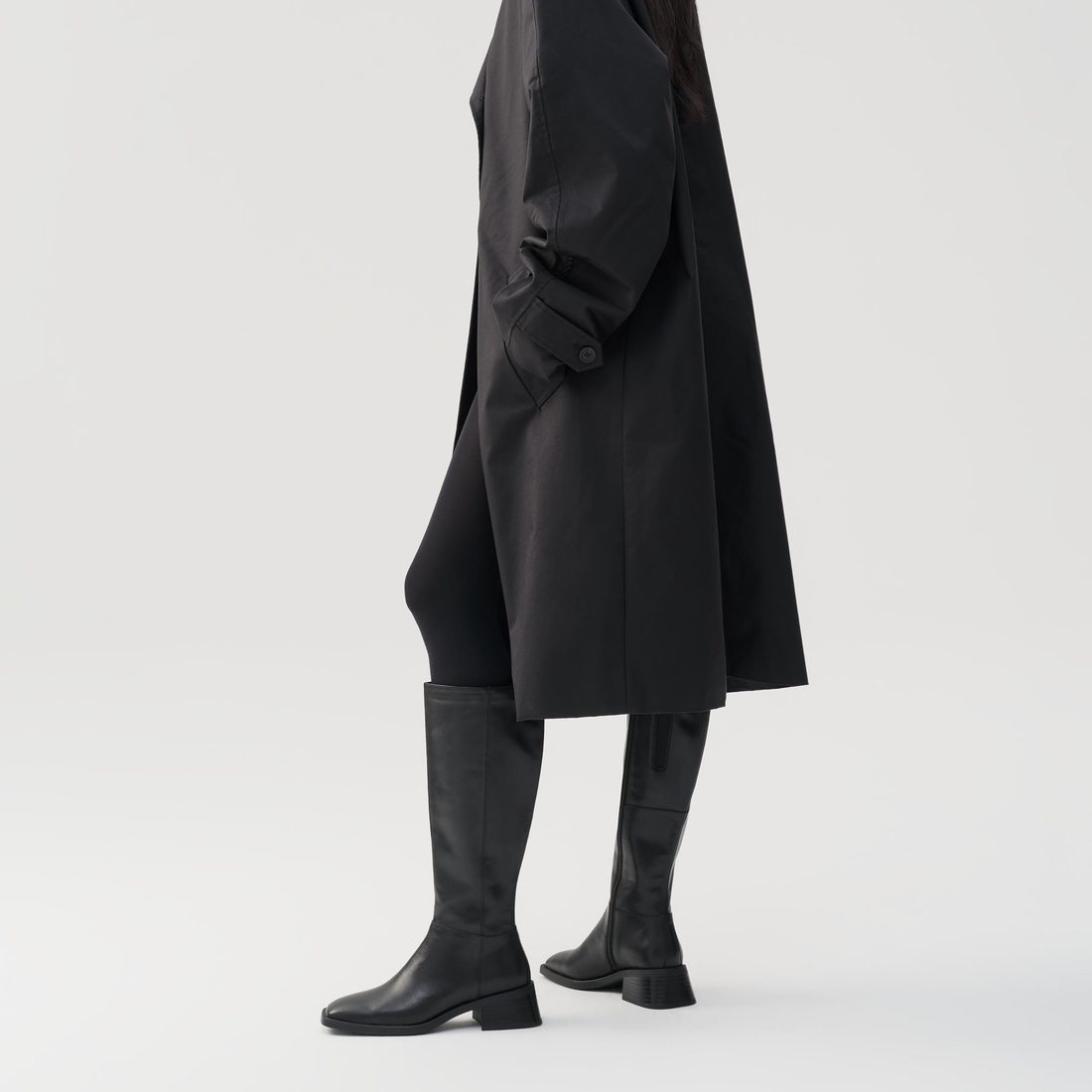 Person wearing a black coat and black knee-high boots on a light gray background