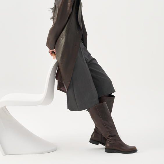Person wearing a long coat and brown knee-high boots sitting on a white chair against a white background